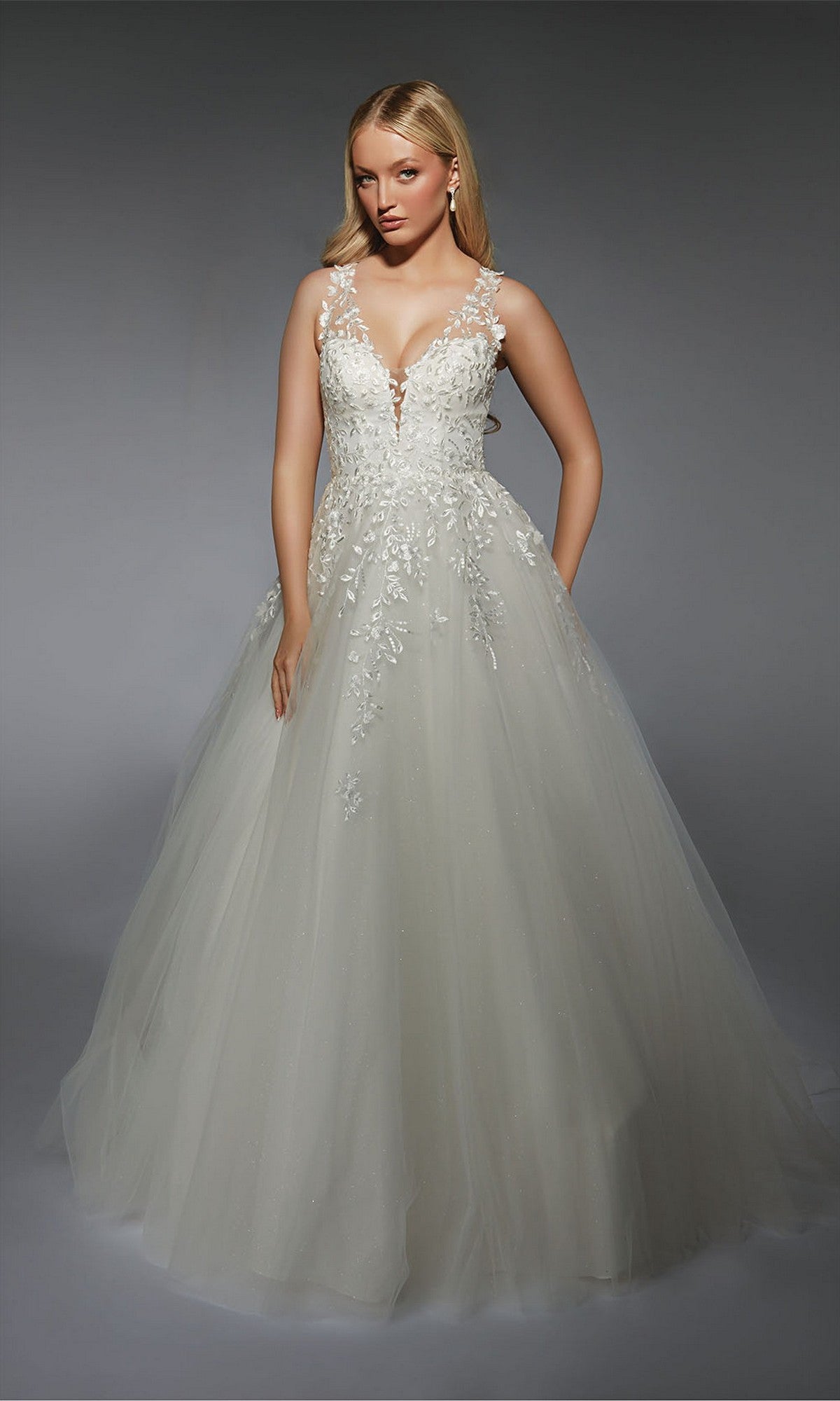 A woman wears the Alyce 7128 long white dress—a bridal ball gown with lace appliqué and floral bodice details, a full tulle skirt, set against a plain gray background.